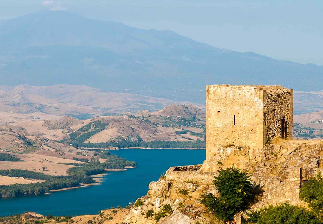 Paesaggio collinare con torre antica e lago, montagne sullo sfondo.