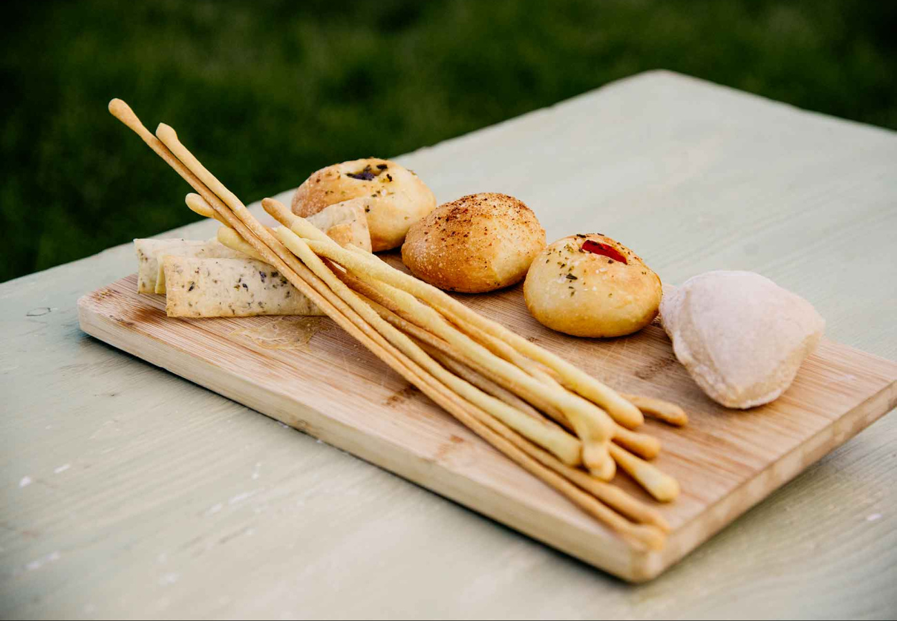Tagliere con grissini, panini aromatizzati e formaggio su tavolo di legno.