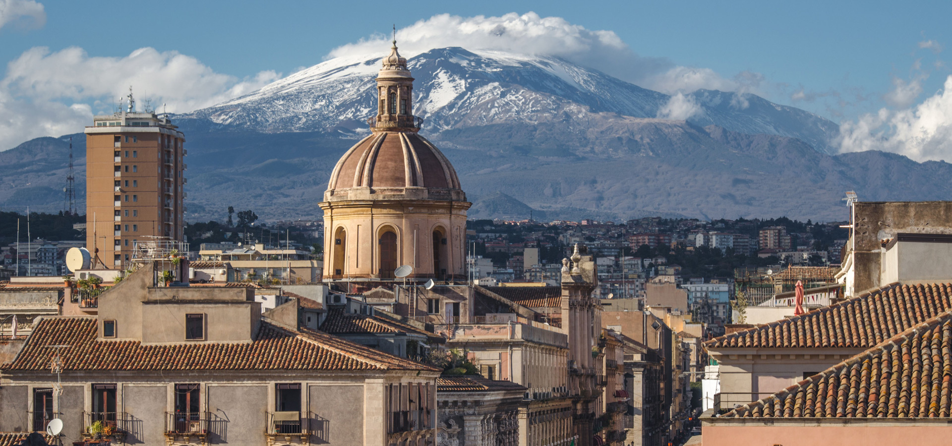Veduta di Catania con l'Etna innevato sullo sfondo e architettura storica in primo piano.