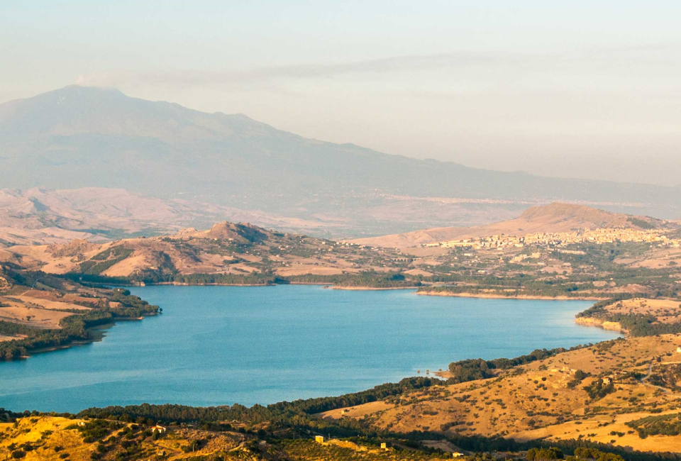Panorama del lago con colline e montagne in lontananza, atmosfera serena e luminosa.